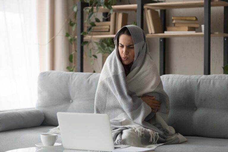 Woman sitting on couch wrapped in blanket looking cold while on computer